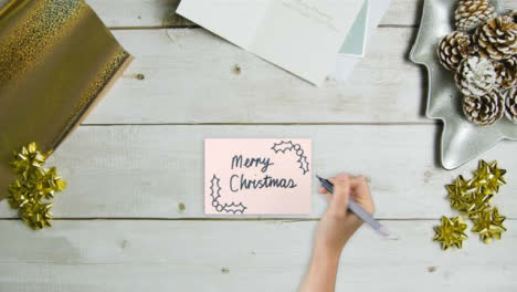 Draufsicht-Der-Hand,-Die-Frohe-Weihnachten-Auf-Papier-Mit-Weihnachtskarten-Und-Geschenken-Schreibt