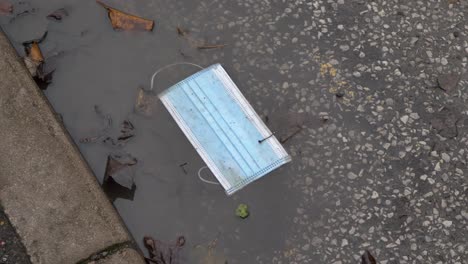 Handheld-High-Angle-Shot-of-Discarded-Face-Mask-In-a-Dirty-Puddle-