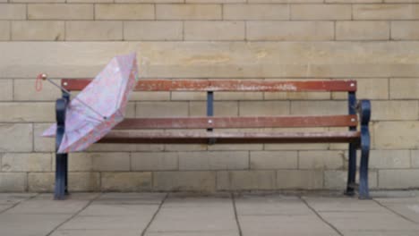 Defocused-Medium-Shot-of-Discarded-Umbrella-On-Bench-as-People-Walk-Past