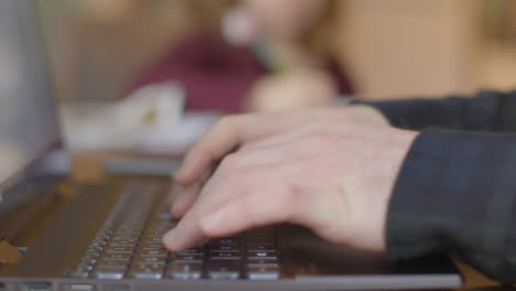 Sliding-Close-Up-Shot-of-Hands-Typing-On-Laptop-with-Child-In-Background-Doing-Homework