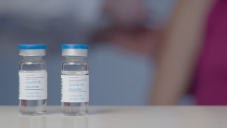 Schieben-Sie-Eine-Nahaufnahme-Von-Covid-Impfstofffläschchen,-Während-Der-Patient-Im-Hintergrund-Eine-Injektion-Erhält