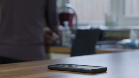 Sliding-Close-Up-Shot-of-Phone-On-Kitchen-Surface-As-Adult-Cleans-In-the-Background