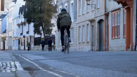 Low-Angle-Shot-of-Cyclist-Riding-Down-Old-Street-Past-Pedestrians-