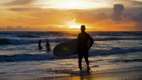 Tiro-Medio-De-Mano-De-Surfista-Caminando-Por-La-Orilla-De-La-Playa-Echo-Siluetas-Contra-La-Puesta-De-Sol