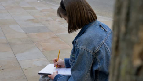 Tracking-Shot-Past-Tree-Revealing-Young-Woman-Sitting-On-Bench-Writing-In-Notebook