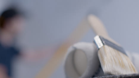 Close-Up-Shot-of-Paint-Brush-and-Roller-with-Person-Painting-In-Background