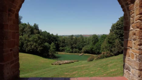 Drone-Shot-Flying-Through-Brick-Arch-to-Reveal-a-Scenic-Lake-On-Edge-of-Woodland