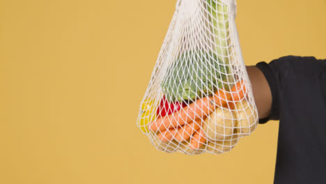 Mid-Shot-of-Man-Holding-up-Bag-of-Vegetables-In-Front-of-Yellow-Background