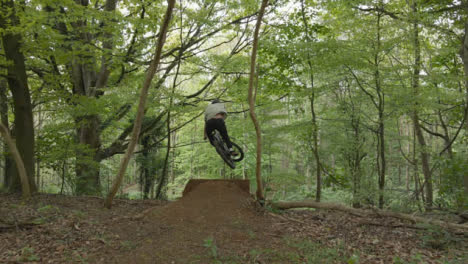 Drone-Tracking-Man-On-Mountain-Bike-Cycling-Doing-Mid-Air-Jump-On-Trail-Through-Countryside-And-Woodland