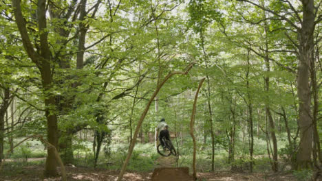 Drone-Tracking-Man-On-Mountain-Bike-Cycling-Doing-Mid-Air-Jumps-On-Trail-Through-Countryside-And-Woodland