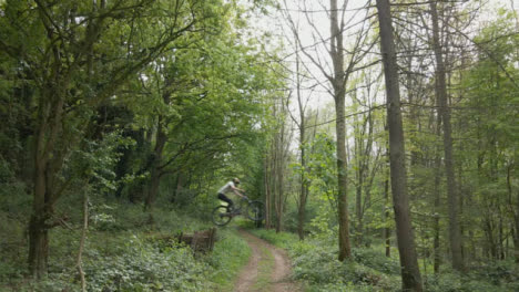 Drone-Tracking-Man-On-Mountain-Bike-Cycling-Doing-Mid-Air-Jump-On-Trail-Through-Countryside-And-Woodland-2