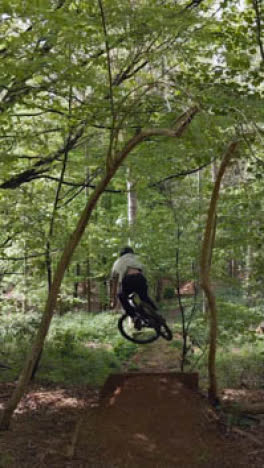 Drone-Vertical-Video-Tracking-Man-On-Mountain-Bike-Cycling-Doing-Mid-Air-Jump-On-Trail-Through-Woodland