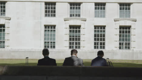 Rear-View-Of-Businessmen-Sitting-On-Bench-With-Suitcases-Outside-London-Office