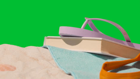 Summer-Holiday-Concept-Of-Book-Flip-Flops-Beach-Towel-On-Sand-Against-Green-Screen