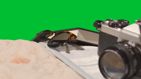 Summer-Holiday-Concept-Of-Sunglasses-Book-Camera-Beach-Towel-On-Sand-Against-Green-Screen