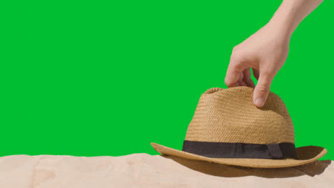 Summer-Holiday-Concept-With-Hand-Picking-Up-Sun-Hat-On-Sand-Against-Green-Screen