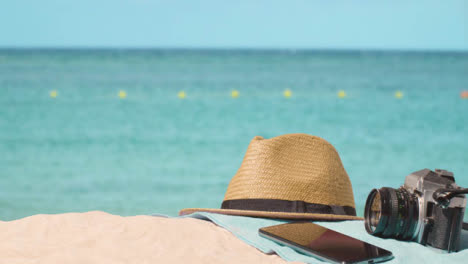 Concepto-De-Vacaciones-De-Verano-De-Cámara-Sombrero-Para-El-Sol-Teléfono-Móvil-Toalla-De-Playa-En-La-Arena-Contra-El-Mar