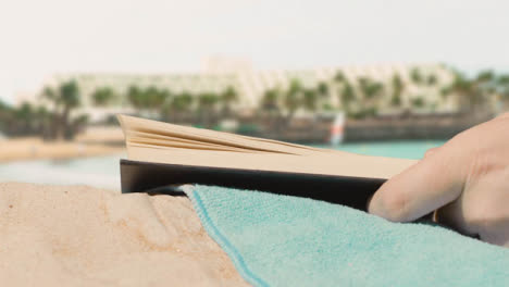 Sommerferienkonzept-Der-Person-Auf-Strandtuch,-Das-Buch-Gegen-Meer--Und-Hotelhintergrund-Liest