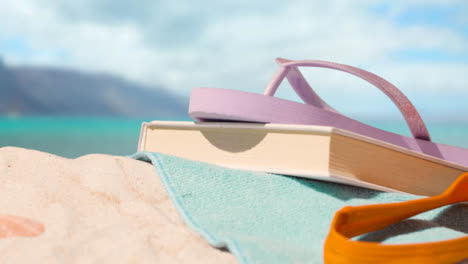 Summer-Holiday-Concept-Of-Book-Flip-Flops-Beach-Towel-On-Sand-Against-Sea-Background