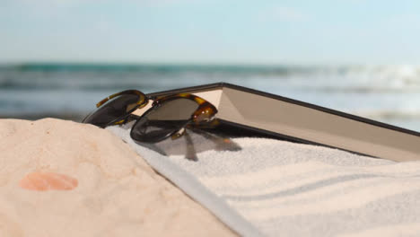 Sommerferienkonzept-Des-Sonnenbrillenbuchbadetuchs-Auf-Sand-Gegen-Seehintergrund