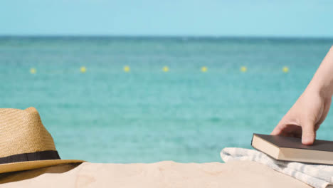 Concepto-De-Vacaciones-De-Verano-De-Libro-Sombrero-Para-El-Sol-Toalla-De-Playa-En-La-Arena-Contra-El-Fondo-Del-Mar