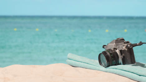 Concepto-De-Vacaciones-De-Verano-De-La-Persona-Que-Recoge-La-Toalla-De-Playa-De-La-Cámara-En-La-Arena-Contra-El-Fondo-Del-Mar