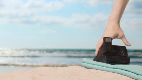 Summer-Holiday-Concept-Of-Person-Picking-Up-Instant-Camera-Beach-Towel-On-Sand-Against-Sea-Background