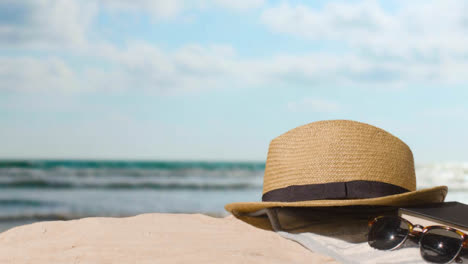 Sommerferienkonzept-Des-Sonnenbrillenbuch-Sonnenhut-Badetuchs-Auf-Sand-Gegen-Seehintergrund