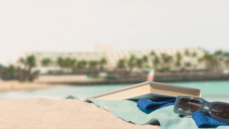 Summer-Holiday-Concept-Of-Person-Picking-Up-Book-On-Beach-Towel-Against-Sea-And-Hotel-Background