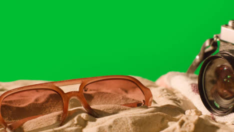 Summer-Holiday-Concept-Of-Sunglasses-Camera-Beach-Towel-On-Sand-Against-Green-Screen