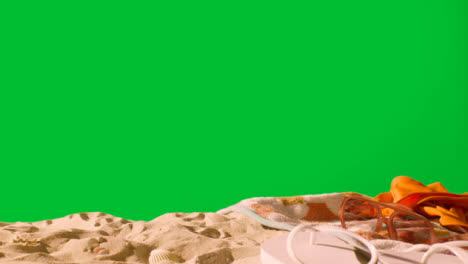 Summer-Holiday-Concept-Of-Sunglasses-Swimsuit-Flip-Flops-Beach-Towel-On-Sand-Against-Green-Screen