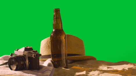 Summer-Holiday-Concept-Of-Beer-Bottle-On-Beach-Towel-With-Camera-And-Sun-Hat-Against-Green-Screen