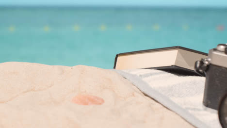 Concepto-De-Vacaciones-De-Verano-De-La-Toalla-De-Playa-De-La-Cámara-Del-Libro-En-La-Arena-Contra-El-Fondo-Del-Mar