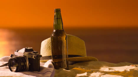 Sommerferienkonzept-Der-Bierflasche-Auf-Strandtuch-Mit-Kamera-Und-Sonnenhut-Gegen-Sonnenunterganghimmel