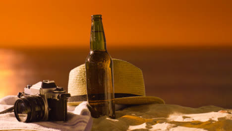 Concepto-De-Vacaciones-De-Verano-De-Botella-De-Cerveza-En-Toalla-De-Playa-Con-Cámara-Y-Sombrero-Para-El-Sol-Contra-El-Cielo-Del-Atardecer