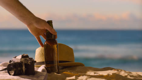 Sommerferienkonzept-Der-Bierflasche-Auf-Strandtuch-Mit-Kamera-Und-Sonnenhut-Gegen-Sonnenunterganghimmel-1