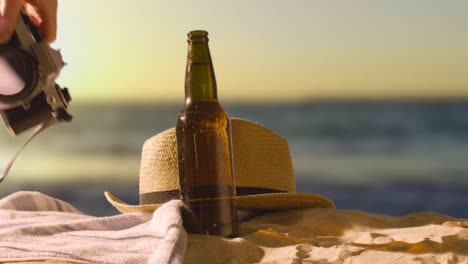 Concepto-De-Vacaciones-De-Verano-De-Botella-De-Cerveza-En-Toalla-De-Playa-Con-Cámara-Y-Sombrero-Para-El-Sol-Contra-El-Cielo-Del-Atardecer-2