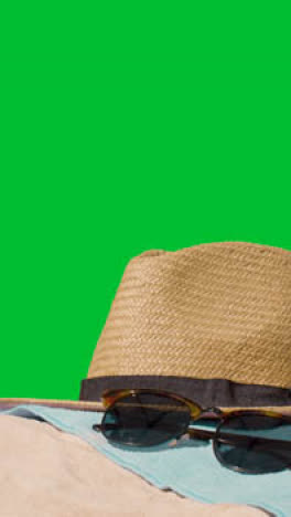 Vertical-Video-Summer-Holiday-Concept-Of-Sunglasses-Sun-Hat-Camera-Beach-Towel-On-Sand-Against-Green-Screen