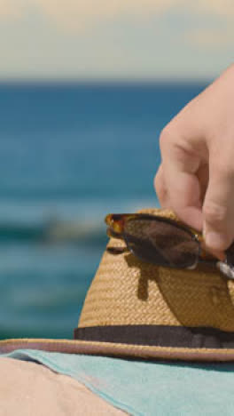 Vertikales-Video-Sommerferienkonzept-Der-Hand,-Die-Sonnenhut-Sonnenbrille-Strandtuch-Auf-Sand-Gegen-Meer-Aufhebt