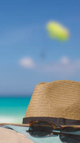 Vertical-Video-Summer-Holiday-Concept-Of-Sunglasses-Sun-Hat-Beach-Towel-On-Sand-Against-Sea-With-Paraglider