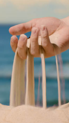 Vertikales-Video-Sommerferienkonzept-Mit-Hand,-Die-Sand-Vom-Strand-Vor-Meereshintergrund-Aufhebt