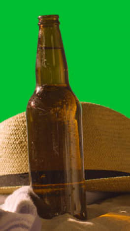 Vertical-Video-Summer-Holiday-Concept-Of-Beer-Bottle-On-Beach-Towel-With-And-Sun-Hat-Against-Green-Screen