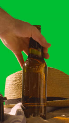 Vertical-Video-Summer-Holiday-Concept-Of-Beer-Bottle-On-Beach-Towel-With-And-Sun-Hat-Against-Green-Screen-1