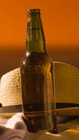 Vertical-Video-Summer-Holiday-Concept-Of-Beer-Bottle-On-Beach-Towel-With-Sun-Hat-Against-Sunset-Sky