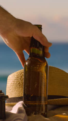 Vertical-Video-Summer-Holiday-Concept-Of-Beer-Bottle-On-Beach-Towel-With-Sun-Hat-Against-Sunset-Sky-1