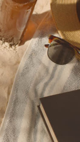 Vertical-Video-Summer-Holiday-Concept-Of-Sunglasses-Book-And-Cold-Beer-On-Beach-Towel-On-Sand