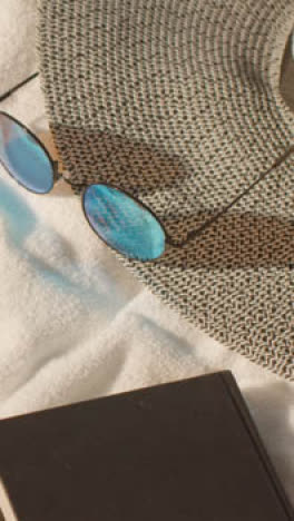 Vertical-Video-Summer-Holiday-Concept-Of-Sunglasses-Sun-Hat-And-Book-On-Beach-Towel-On-Sand