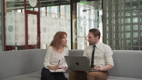 A-Red-Haired-Businesswoman-Has-A-Relaxed-Meeting-With-A-Young-Man-With-A-Laptop-On-The-Couch-In-The-Common-Areas-Of-The-Office-Building-1