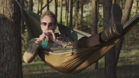 A-Relaxed-Guy-Takes-A-Drink-Lying-In-A-Hammock-In-The-Middle-Of-The-Forest
