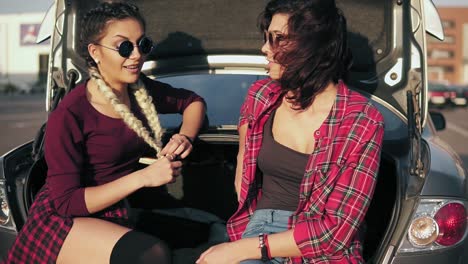 Dos-Jóvenes-Mujeres-Atractivas-Con-Elegantes-Gafas-De-Sol-Sentadas-En-El-Baúl-Del-Camión-En-El-Estacionamiento-Junto-Al-Centro-Comercial-Y-Hablando-Durante-El-Día-Soleado